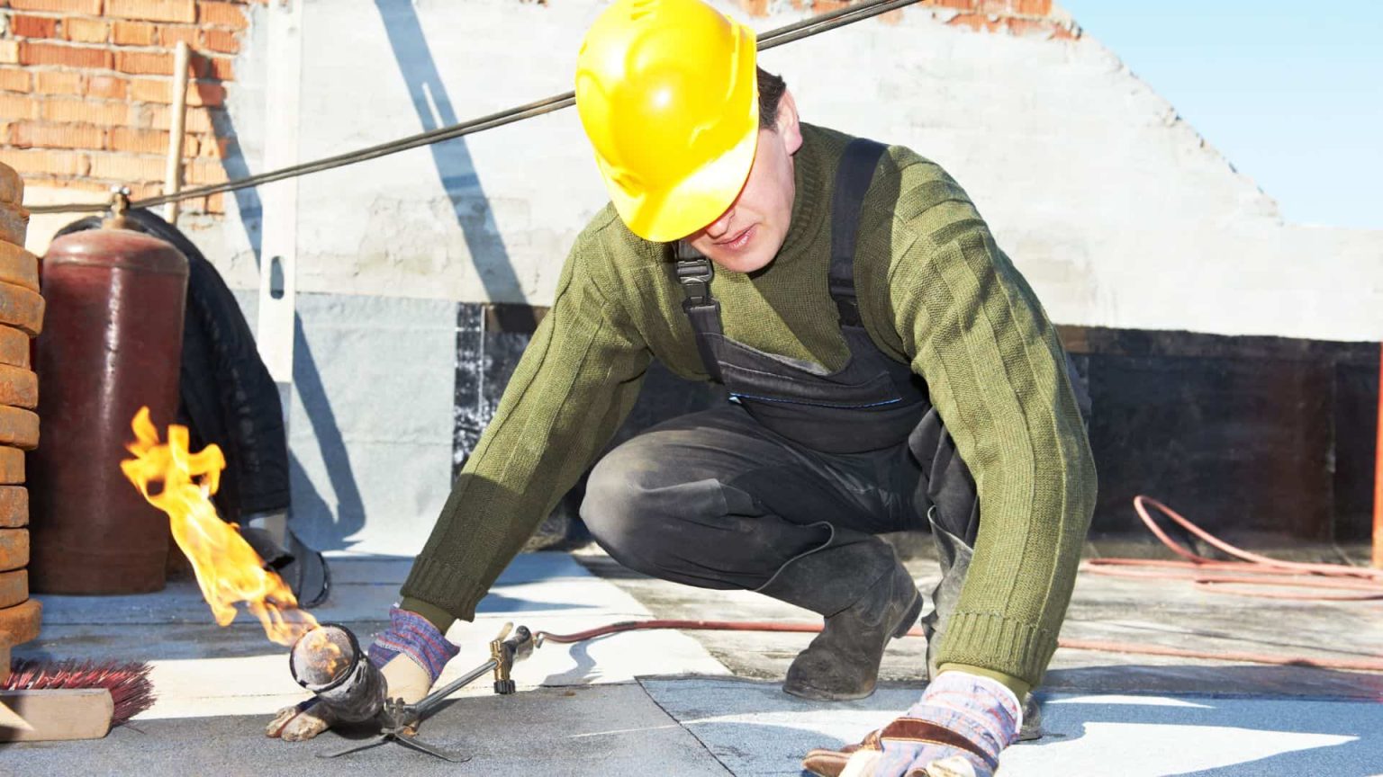 Worker using a torch to install roofing materials, wearing a yellow hard hat and gloves, on a construction site with bricks and equipment in the background.