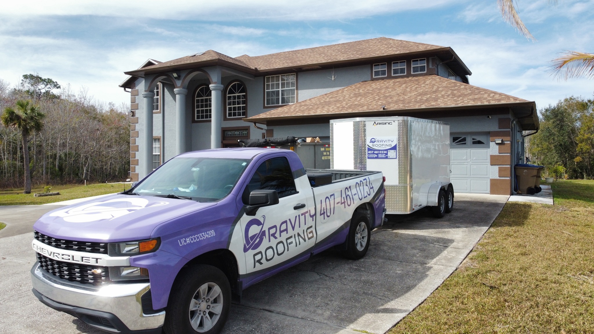 Gravity Roofing truck parked in front of a residential home, showcasing the company's branding and contact information, with a trailer and a well-maintained lawn in the background.