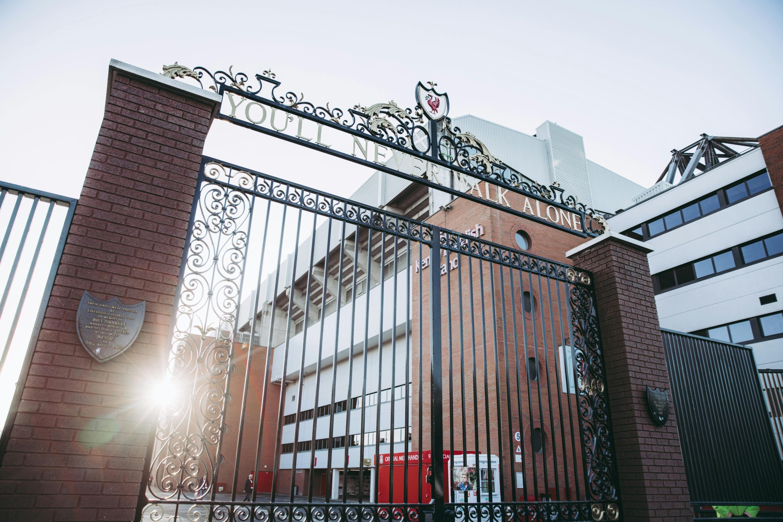 Ornate black gate with "YOU'LL NEVER WALK ALONE" inscription, leading to a stadium, sunlight shining through, symbolizing community and support in sports.