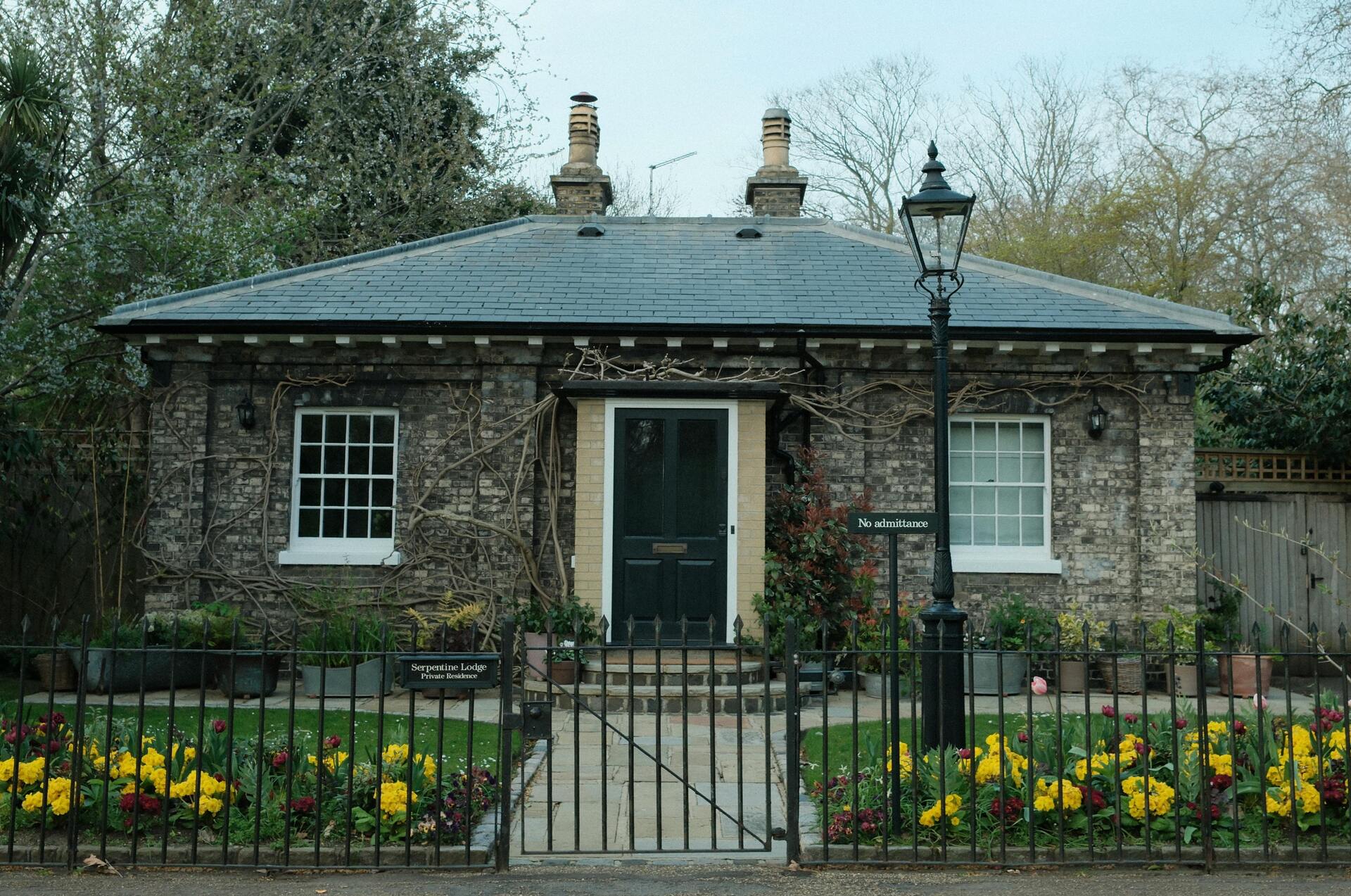 Charming brick house with a dark green door, surrounded by a black iron gate and vibrant flower beds, featuring a "No admittance" sign, emphasizing secure residential aesthetics.