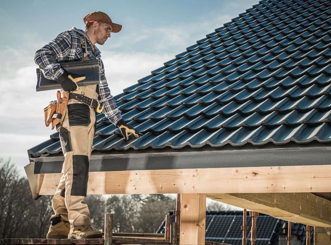 Construction worker inspecting a new roof with blue tiles, wearing safety gloves and holding tools, illustrating roofing services relevant to home repair and maintenance.