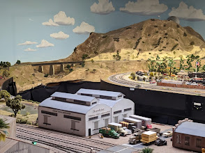 Model train landscape featuring a warehouse, trucks, and mountainous terrain, representing a miniature scene relevant to local attractions in Corona, California.