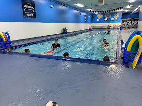 Indoor swimming pool with children and adults engaged in swimming lessons, featuring colorful pool floats and a bright blue environment.
