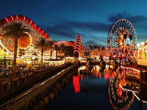 Vibrant night scene at Disney California Adventure Park featuring illuminated attractions like a Ferris wheel and roller coaster, reflecting in the water, surrounded by palm trees.