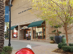 Exterior view of a shopping center featuring a bridal exchange storefront with green awnings, surrounded by palm trees and decorative planters, located in Corona, California, near Joe's Fencing & Gates services.