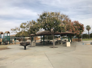 Park pavilion with tables and grill surrounded by trees and playground equipment in Corona, California.