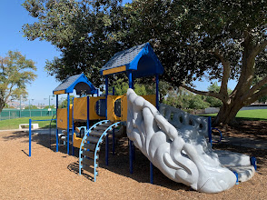 Playground equipment featuring a blue and yellow structure with a slide and climbing elements, surrounded by trees and open green space, suitable for family-friendly community parks in Corona, California.